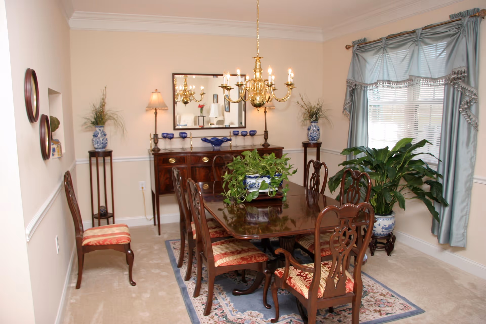 A traditional dining room with a polished wooden dining table surrounded by six chairs with red patterned cushions. The table has a green potted plant centerpiece. The room features beige walls with white trim, a large window with blue curtains, two tall plant stands with blue and white vases holding plants, a wooden sideboard with decorative items, a mirror above it, and a brass chandelier hanging from the ceiling.