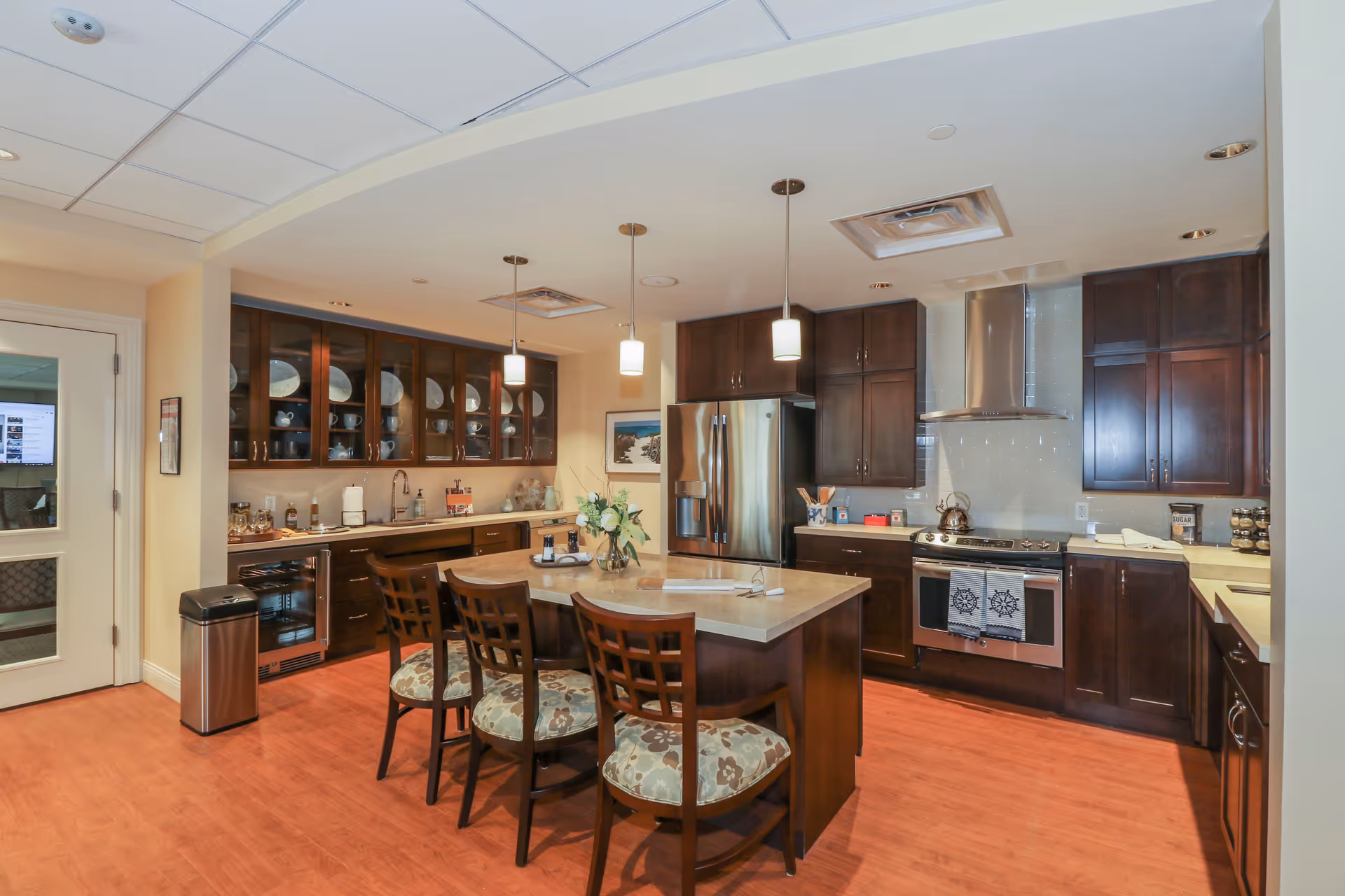 A spacious kitchen with dark wood cabinets, a large island with a beige countertop, and four wooden chairs with patterned cushions. The kitchen features stainless steel appliances including a refrigerator and a stove with a modern range hood. There are pendant lights hanging above the island, glass-front cabinets displaying dishes, and a trash can near a door with a window showing a room beyond.
