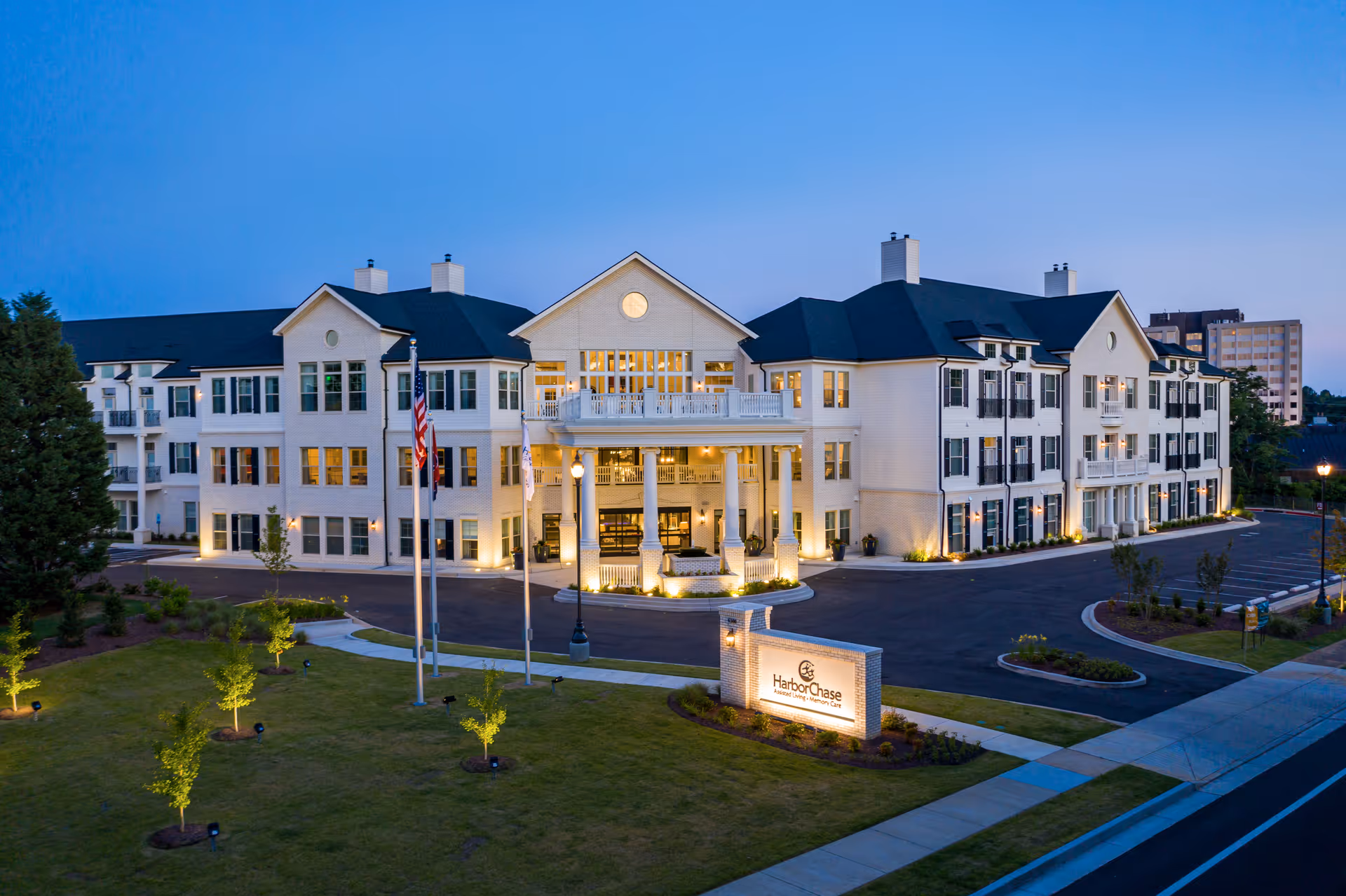 Exterior view of a large, well-lit senior living facility building at dusk with a manicured lawn, flagpoles, and a sign that reads HarborChase Assisted Living & Memory Care.