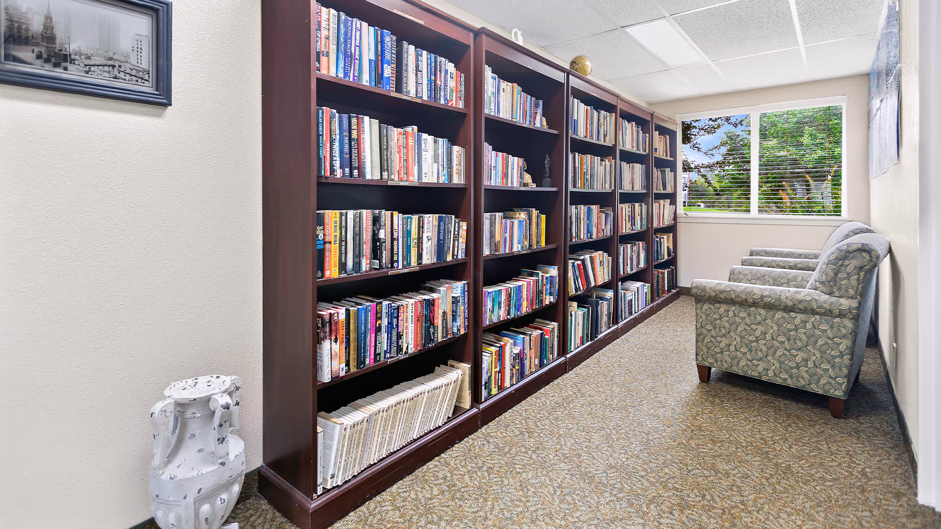 A quiet reading area in Colonial Village featuring tall wooden bookshelves filled with books along one wall, a window with a view of green trees, and two patterned upholstered armchairs positioned against the opposite wall.