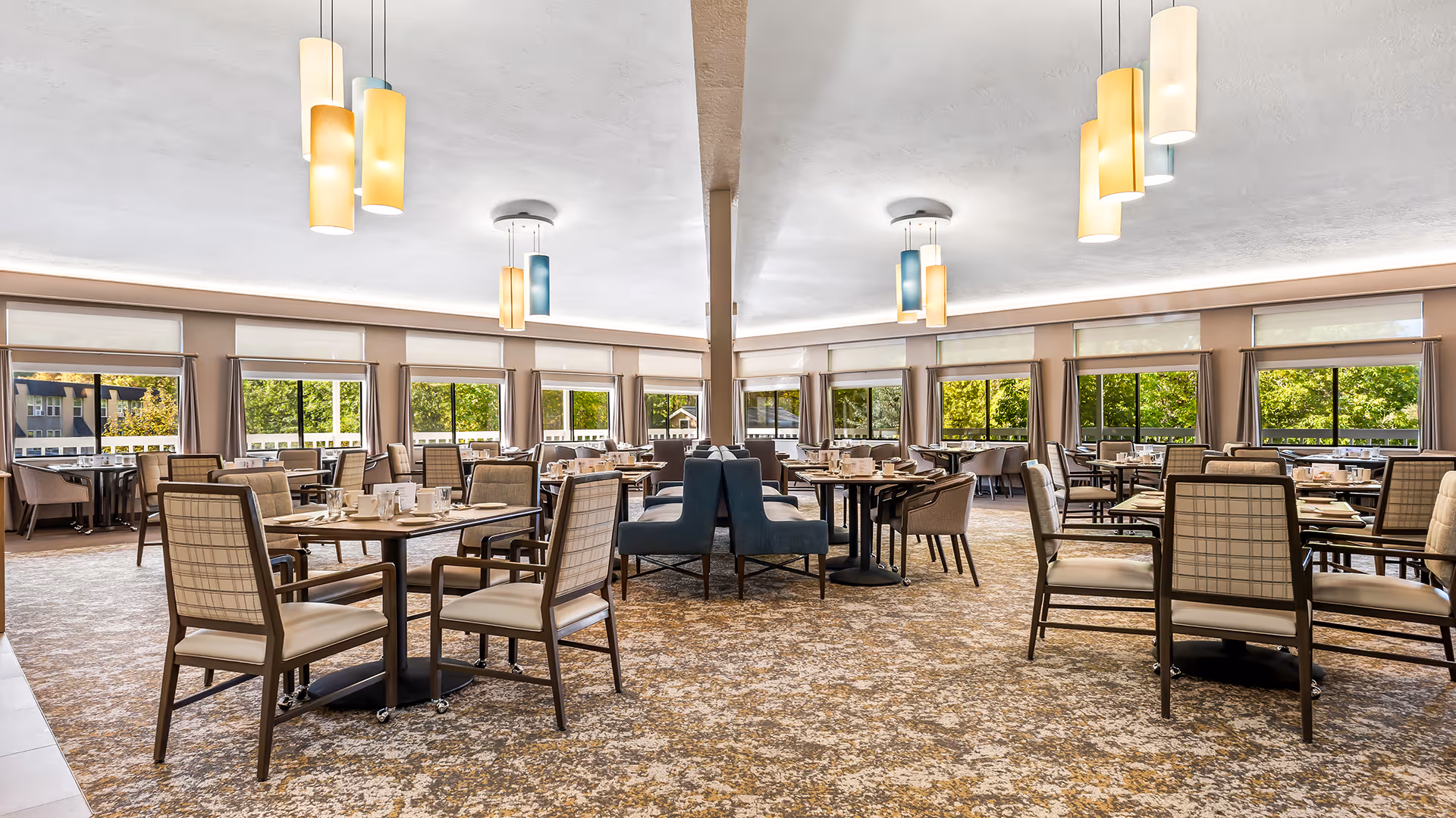 A spacious and well-lit dining room with multiple tables and chairs arranged neatly. The room features large windows with views of greenery outside, beige walls, patterned carpet flooring, and modern pendant lights hanging from the ceiling. Tables are set with plates, cups, and utensils, ready for dining.