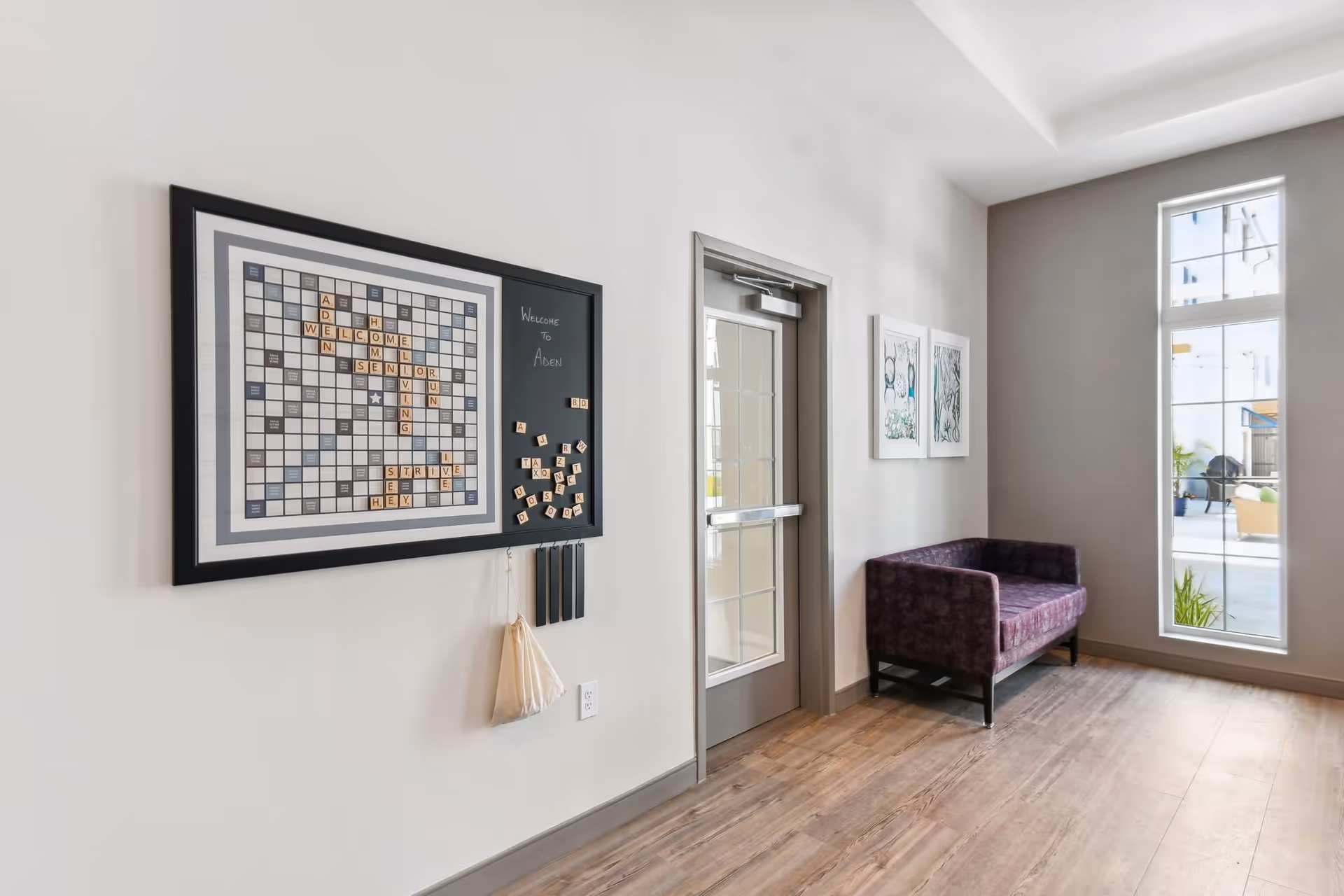 A bright hallway in a senior living facility with a large Scrabble-style magnetic board on the wall displaying the words 'Aden Senior Living Welcome'. Next to the board is a door with glass panels, and a purple upholstered bench is placed against the wall near a tall window letting in natural light. Two framed artworks hang on the wall above the bench.