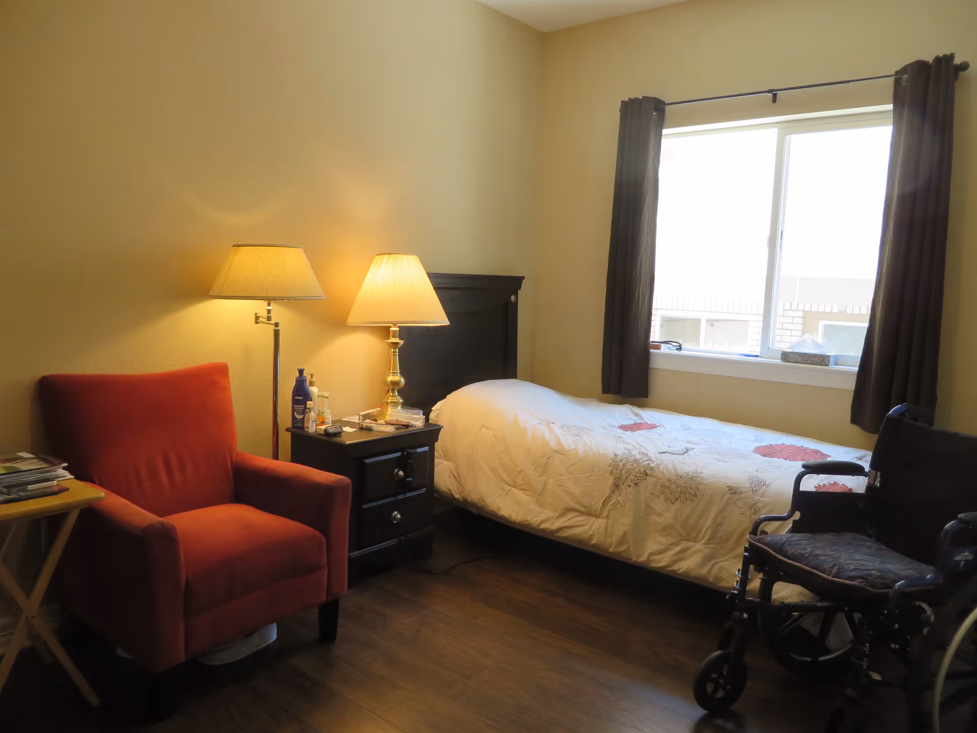 A small bedroom with a single bed covered with a white floral comforter, a black wheelchair next to the bed, a black nightstand with two lamps and various personal items, a red armchair, and a window with dark curtains letting in natural light.