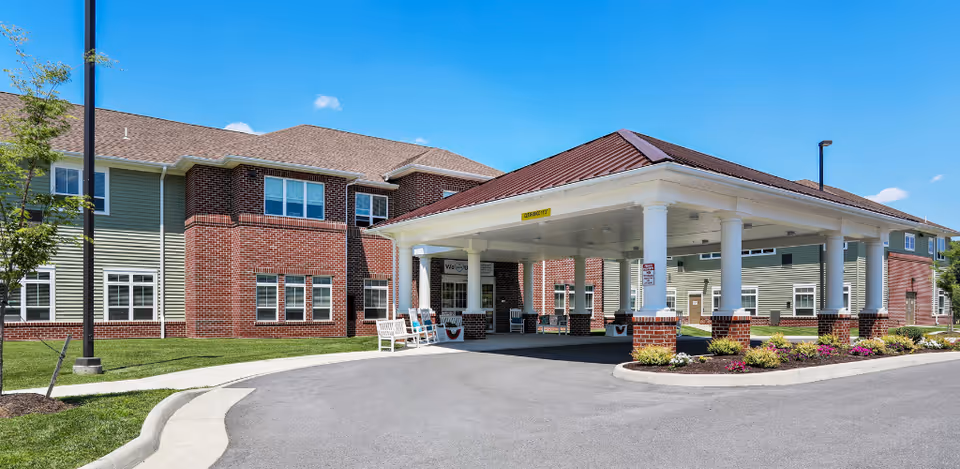 Covered entrance and porte-cochère of a two-story brick and siding senior living building with benches, landscaping, and a driveway.