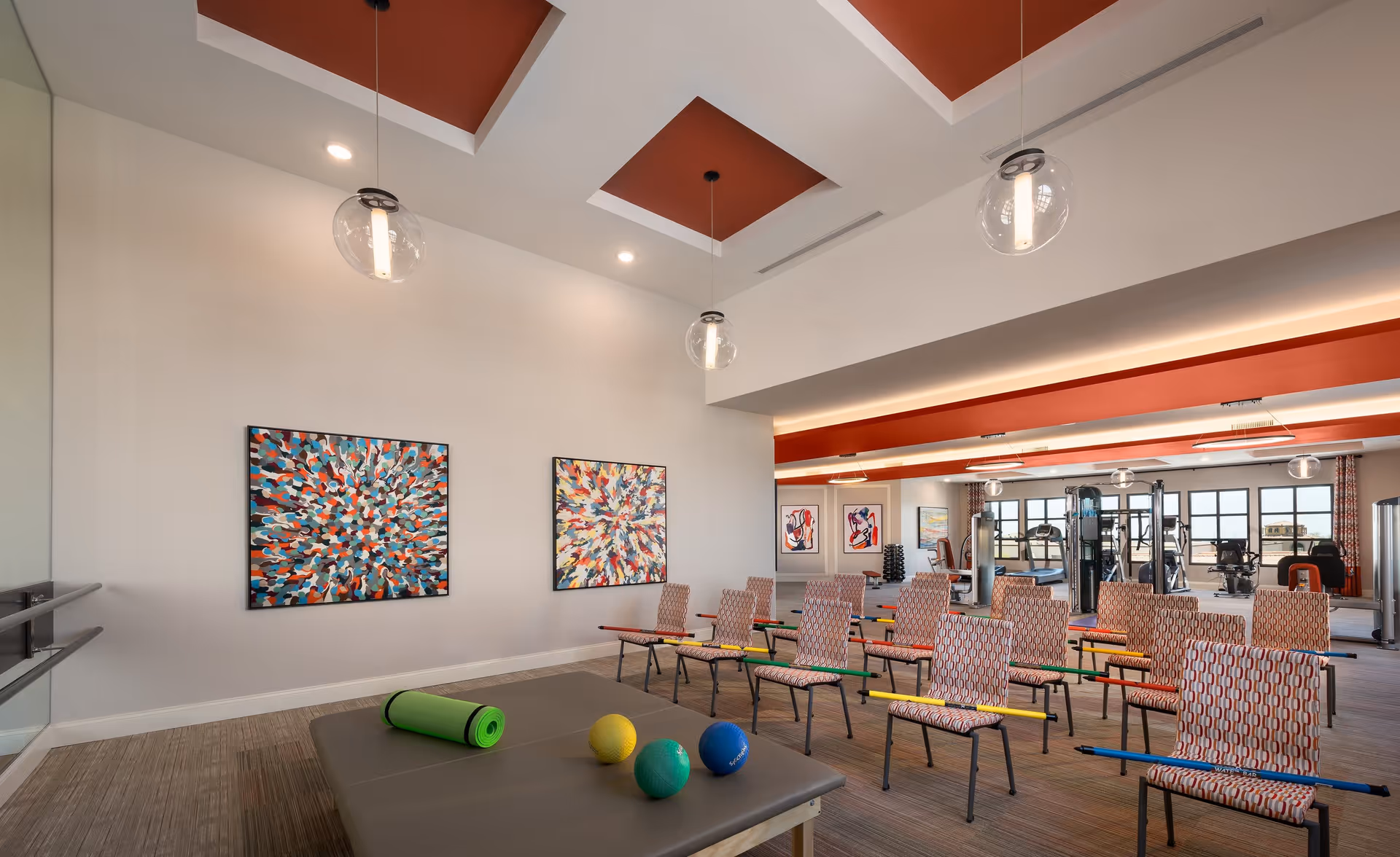 A spacious exercise room with rows of chairs equipped with colorful exercise bars. The room features modern ceiling lights, two abstract paintings on the wall, and a mirrored wall on the left side. In the foreground, there is a padded table with a green yoga mat and three small exercise balls. In the background, gym equipment and large windows letting in natural light are visible.