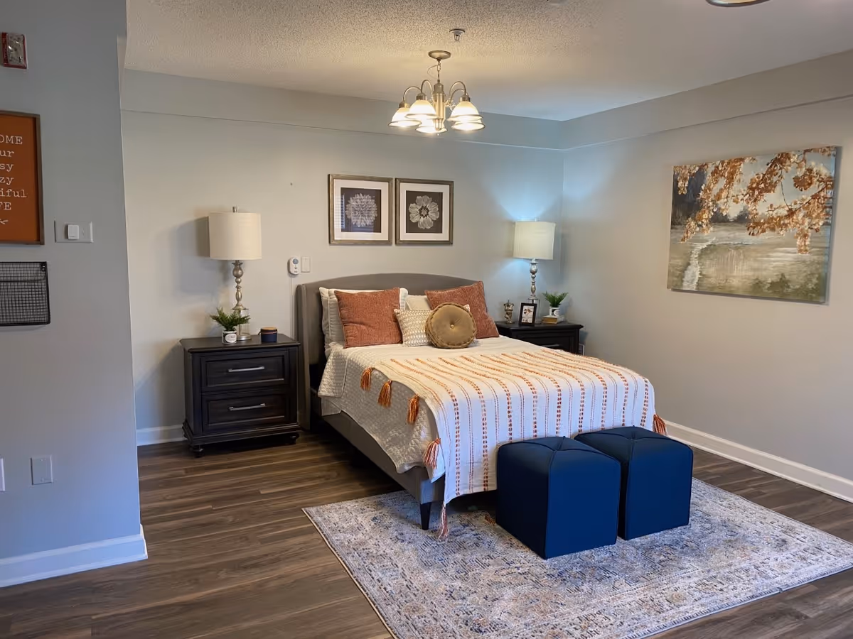 A neatly arranged bedroom with a bed centered against a light gray wall. The bed has a gray upholstered headboard, white bedding with a striped throw blanket, and several decorative pillows in shades of orange and beige. On either side of the bed are dark wooden nightstands with matching lamps, small plants, and decorative items. Above the bed are two framed floral prints, and on the right wall is a large painting of a tree with autumn leaves over a body of water. The floor is wooden with a patterned area rug under the bed and two dark blue ottomans at the foot of the bed.