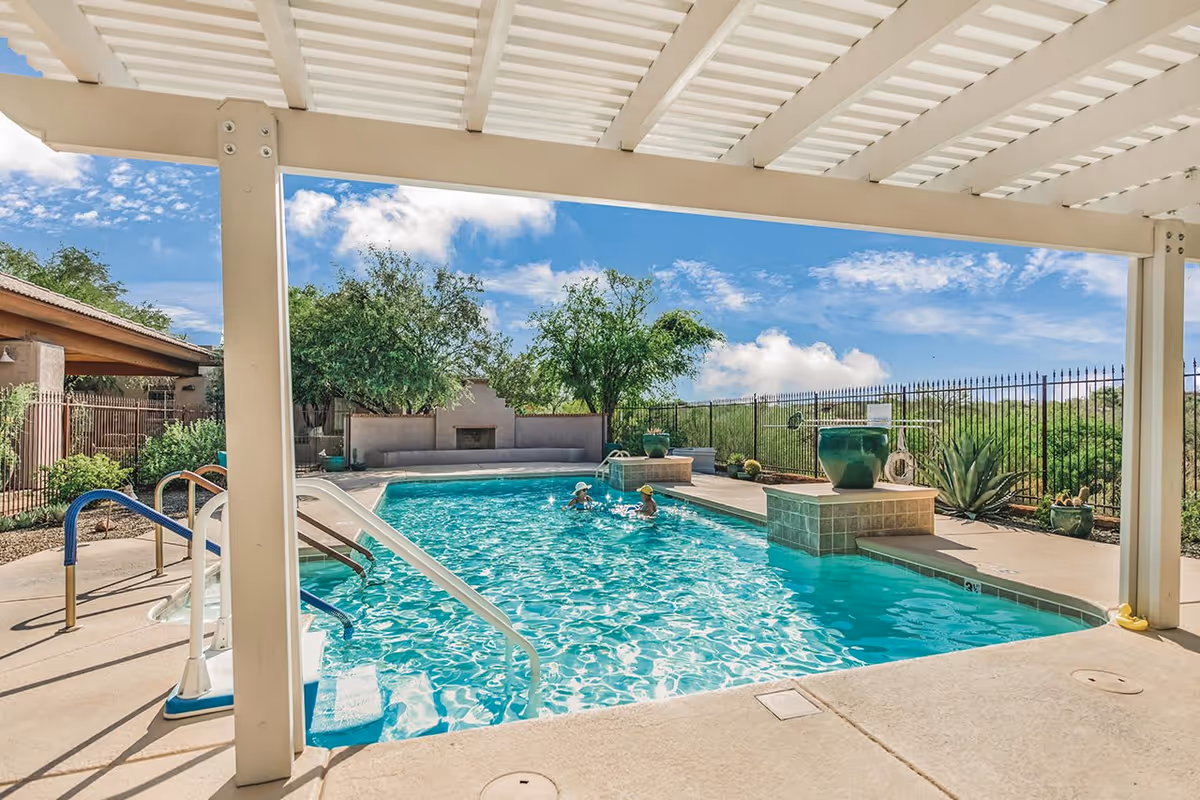 Outdoor swimming pool area with two people in the water, surrounded by a concrete deck and greenery. The pool is partially shaded by a white pergola, and there is a metal handrail and pool steps visible. The background includes trees, a fence, and a clear blue sky with some clouds.
