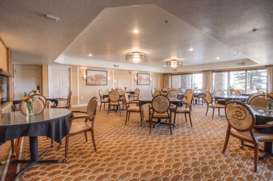 A spacious dining room with multiple round tables covered with black tablecloths, each surrounded by elegant chairs with patterned upholstery. Small flower arrangements are placed on each table. The room has large windows with beige curtains allowing natural light to enter, and warm lighting fixtures on the ceiling. The walls are decorated with framed landscape paintings.