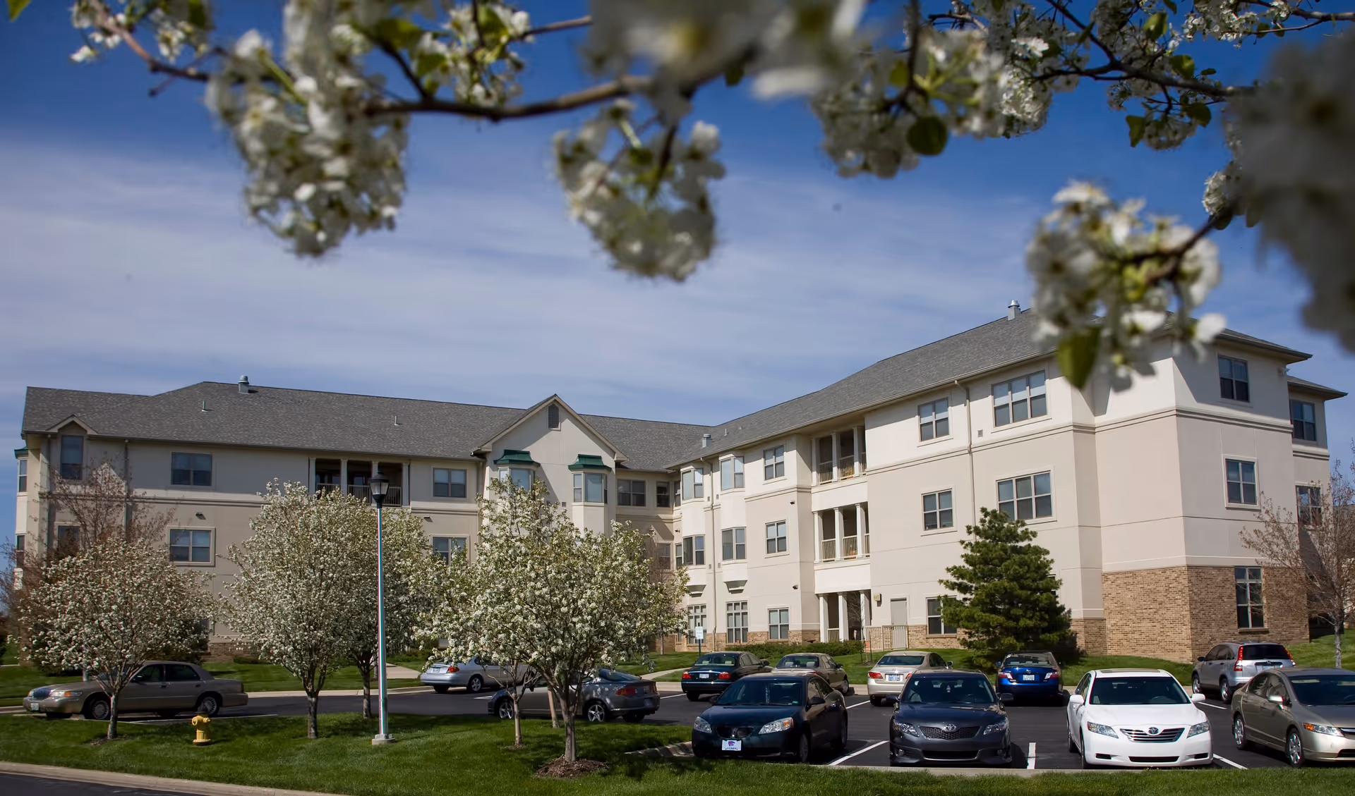 Exterior view of a three-story senior living facility building with beige and light brown walls, surrounded by a parking lot with several parked cars and blooming trees in the foreground under a clear blue sky.
