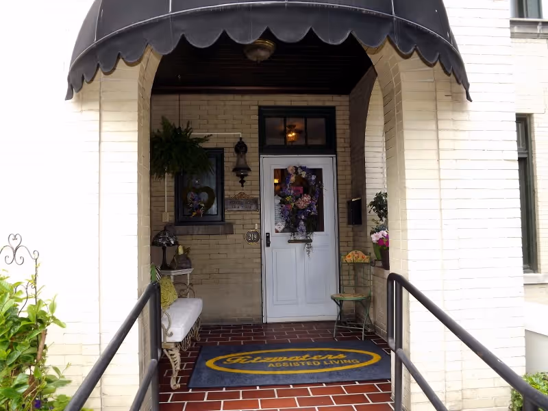 Entrance to Fitzwater's Assisted Living with a white door decorated with a floral wreath, a black awning overhead, a welcome mat with the facility's name, a bench with cushions on the left, and a small chair with potted flowers on the right.