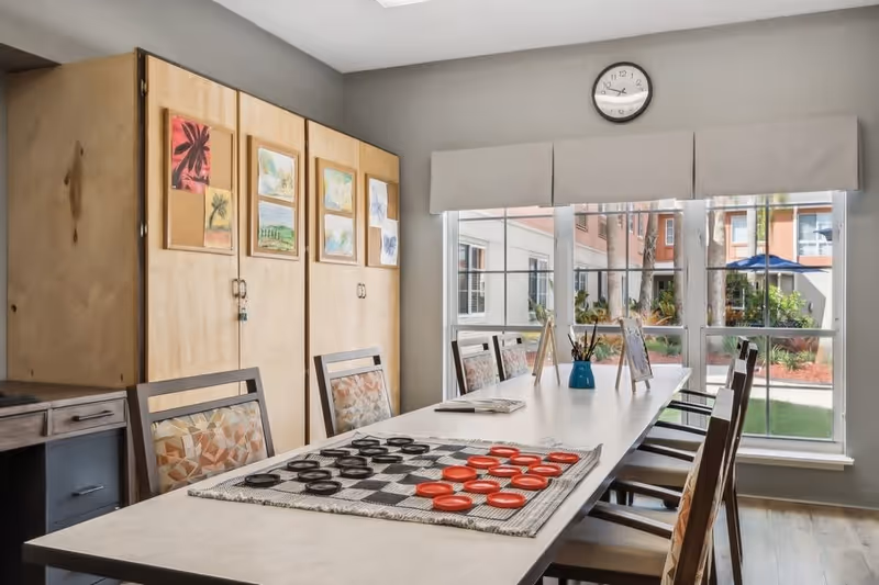 A bright dining room featuring a large table with a checkers game set up, surrounded by chairs. The walls are adorned with colorful artwork, and large windows provide a view of the outdoor area.