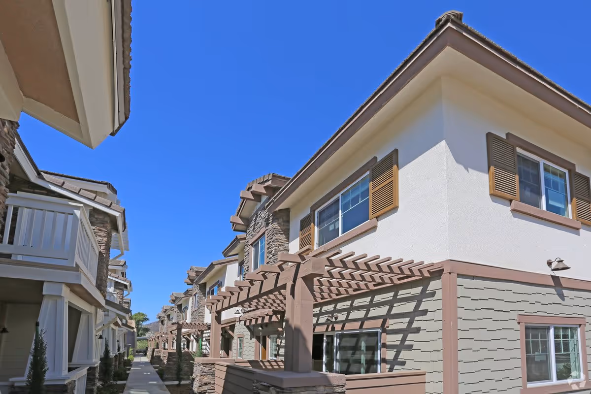 Exterior view of a modern residential building complex with two-story units featuring balconies, pergolas, and a clear blue sky above.