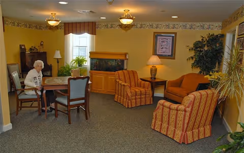 A cozy living room area with yellow walls and carpeted floor. The room features a wooden table with four chairs, one of which is occupied by an elderly woman. There are two patterned armchairs and a matching sofa arranged around a small wooden side table with a lamp. A large fish tank is set against the wall beneath a window with a valance. Various plants and framed artwork decorate the room.