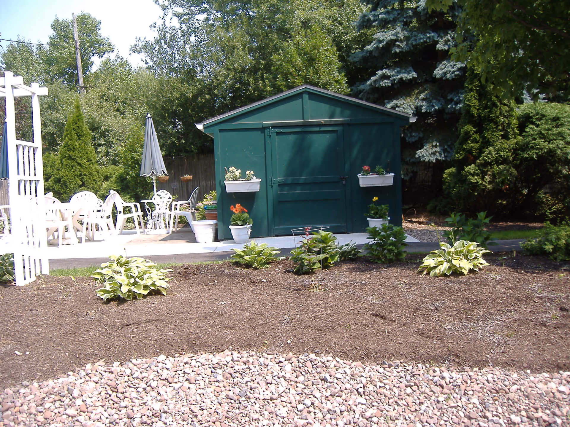 Outdoor garden area with a green shed in the center, decorated with flower pots on either side of the door. In front of the shed is a mulched garden bed with small plants. To the left, there is a patio area with white outdoor chairs and tables, and a closed umbrella. Trees and shrubs surround the area.