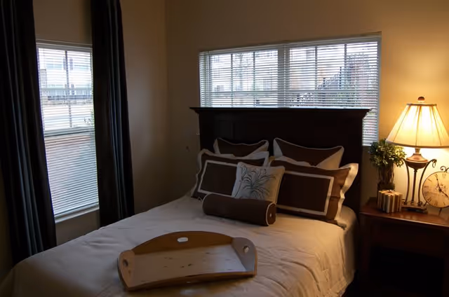 A softly lit bedroom with a made bed topped with brown and white pillows, a wooden headboard, a nightstand with a lamp and clock, and windows with blinds.