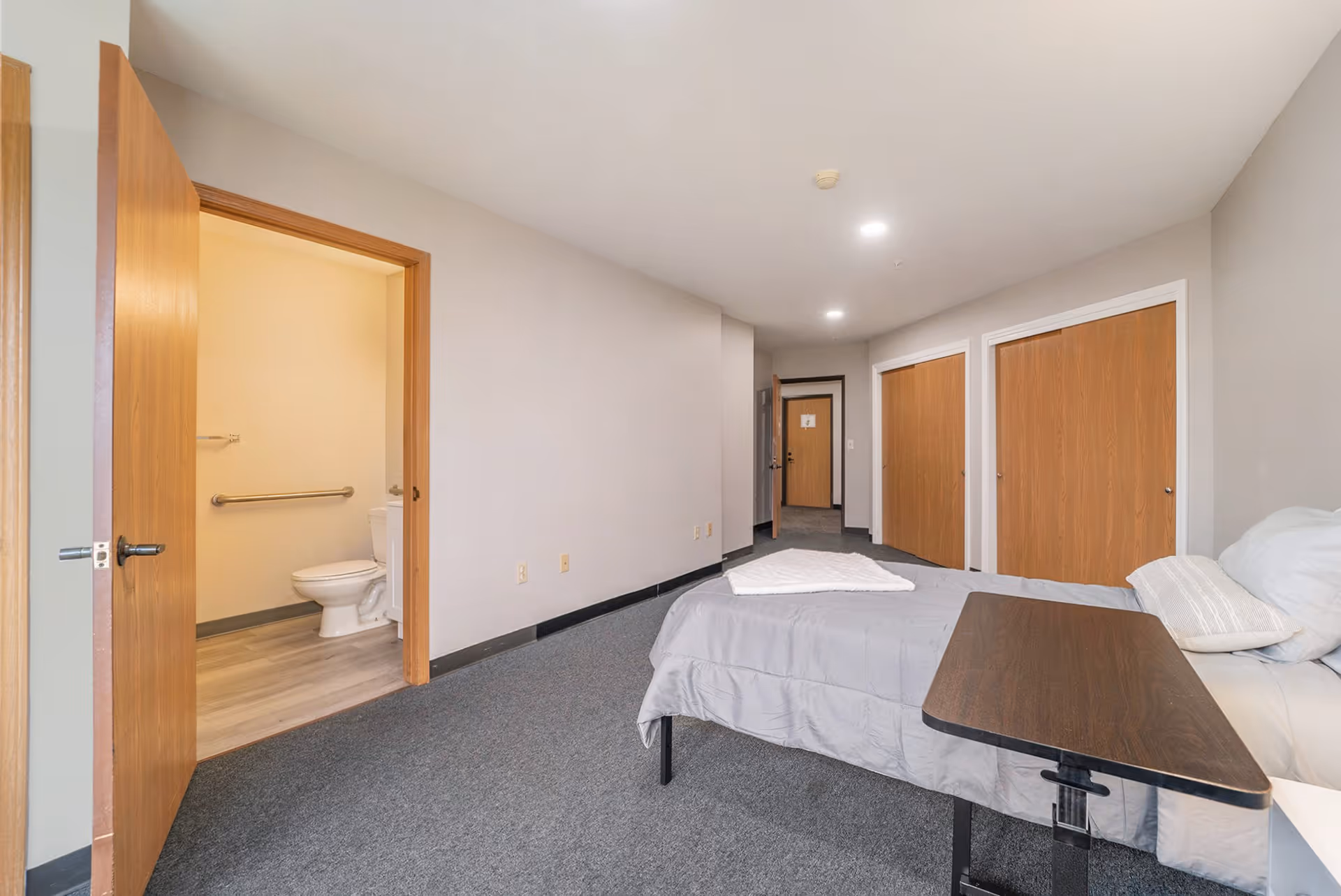 A simple bedroom in a senior living facility with a single bed covered in gray bedding and a pillow. There is a wooden overbed table on wheels next to the bed. The room has gray carpet flooring and light gray walls. Two closed wooden closet doors are visible on the right side of the room. An open door on the left side reveals a bathroom with a toilet and grab bars. The hallway and entrance door are visible in the background.