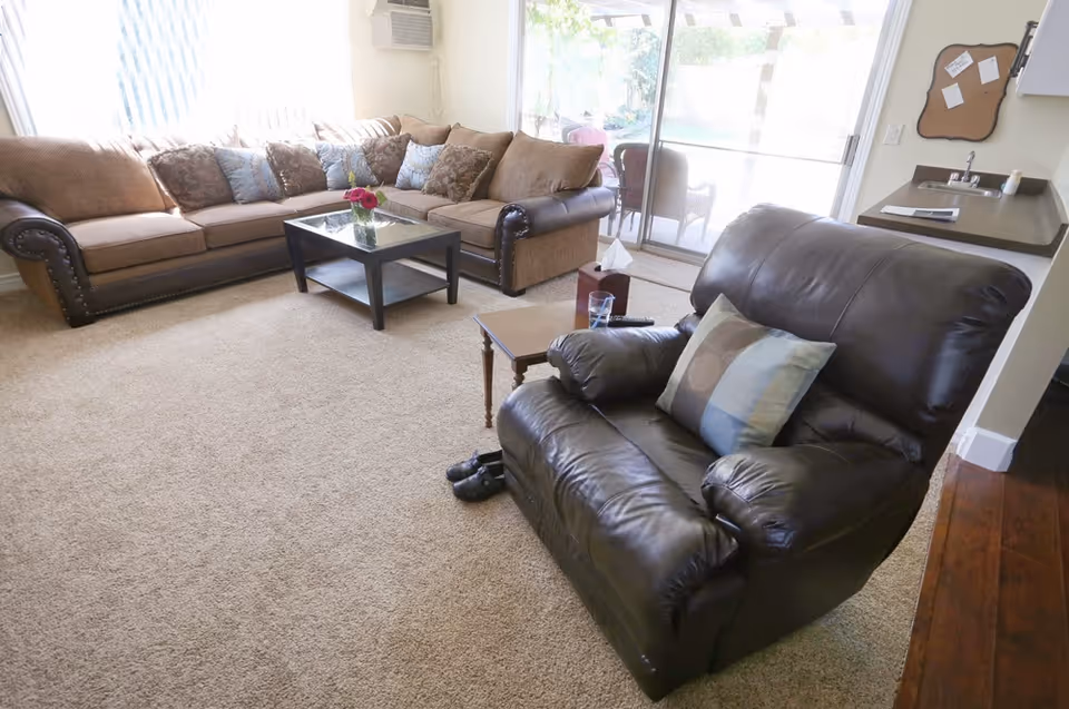 A cozy living room with a large brown sectional sofa adorned with multiple patterned cushions, a black leather armchair with a decorative pillow, a glass-top coffee table with a vase of flowers, and a small side table holding a glass of water and a remote control. The room has beige carpeting, a sliding glass door leading to an outdoor patio, and a small kitchen counter with a sink in the background.