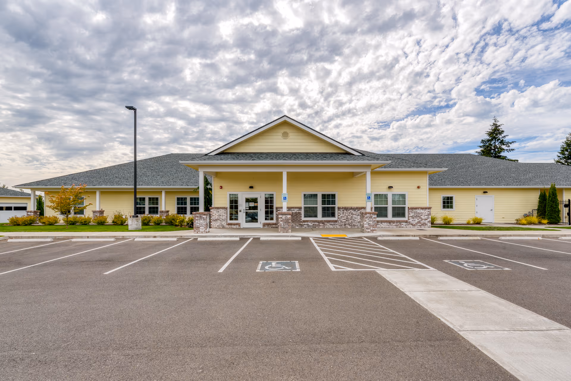 Front exterior view of Easthaven Villa Assisted Living & Memory Care building with a yellow facade, brick accents, multiple windows, a covered entrance, and an empty parking lot with designated handicapped parking spaces under a partly cloudy sky.