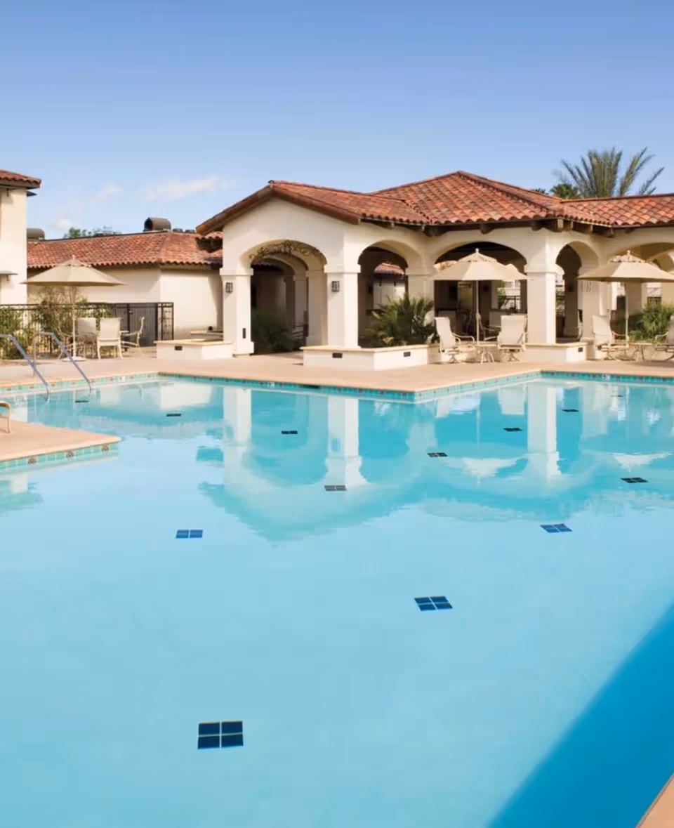 Outdoor swimming pool with lounge chairs, umbrellas and a Mediterranean-style clubhouse reflected in the water.