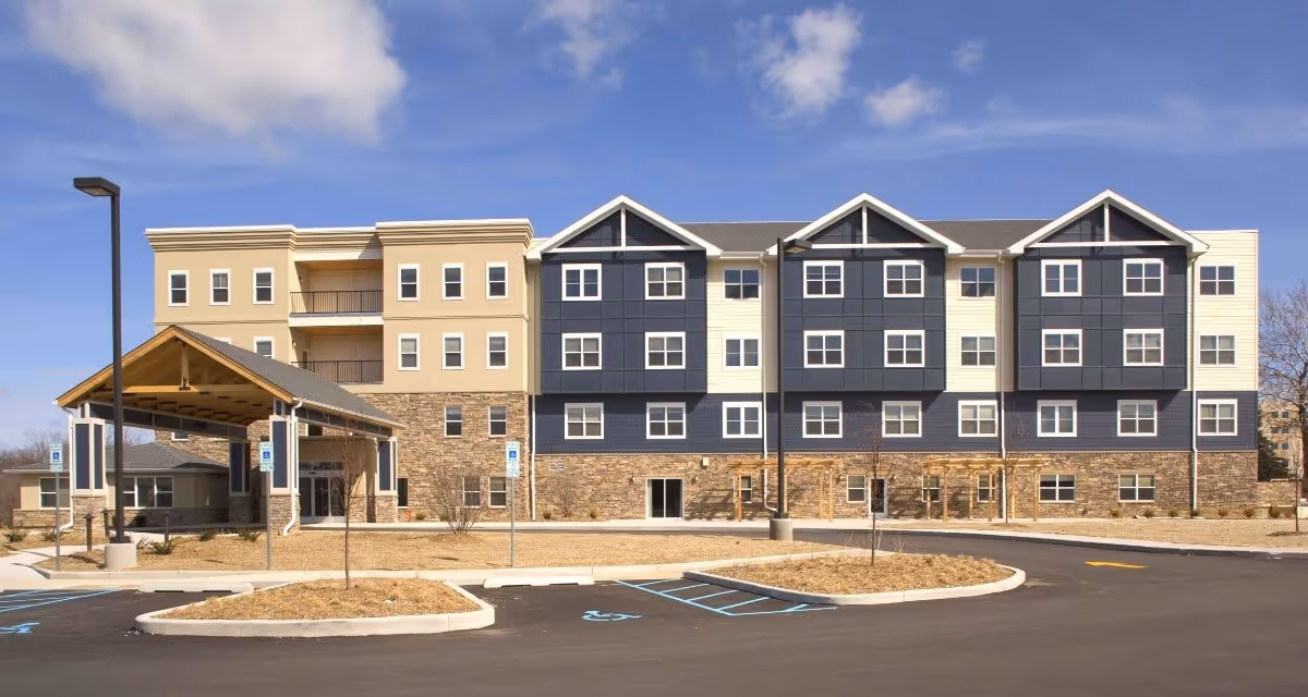 Exterior view of a multi-story senior living facility building with a covered entrance, multiple windows, and a parking lot with handicap parking spaces in front. The building has a combination of beige, blue, and stone facade under a partly cloudy sky.