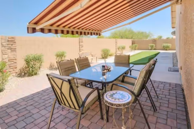 Outdoor patio area with a glass-top table surrounded by six cushioned chairs on a brick-paved floor. There is a small round side table with a decorative top next to one of the chairs. Above the seating area is a striped retractable awning providing shade. In the background, there is a low wall with some small bushes and a green putting green on a concrete path.