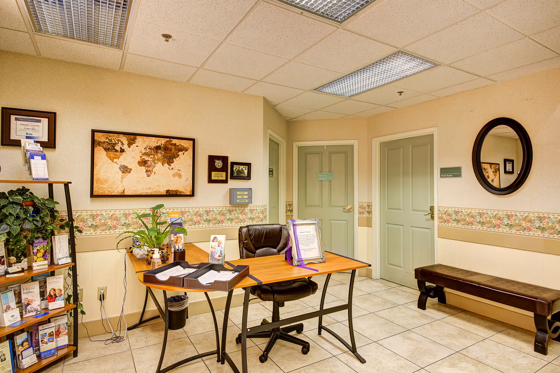 A small office area in an assisted living facility with a wooden desk, black office chair, and various informational brochures on a nearby shelf. The walls are decorated with a world map, plaques, and a round mirror. There are two closed doors and a bench along the wall, with floral wallpaper trim and tiled flooring.