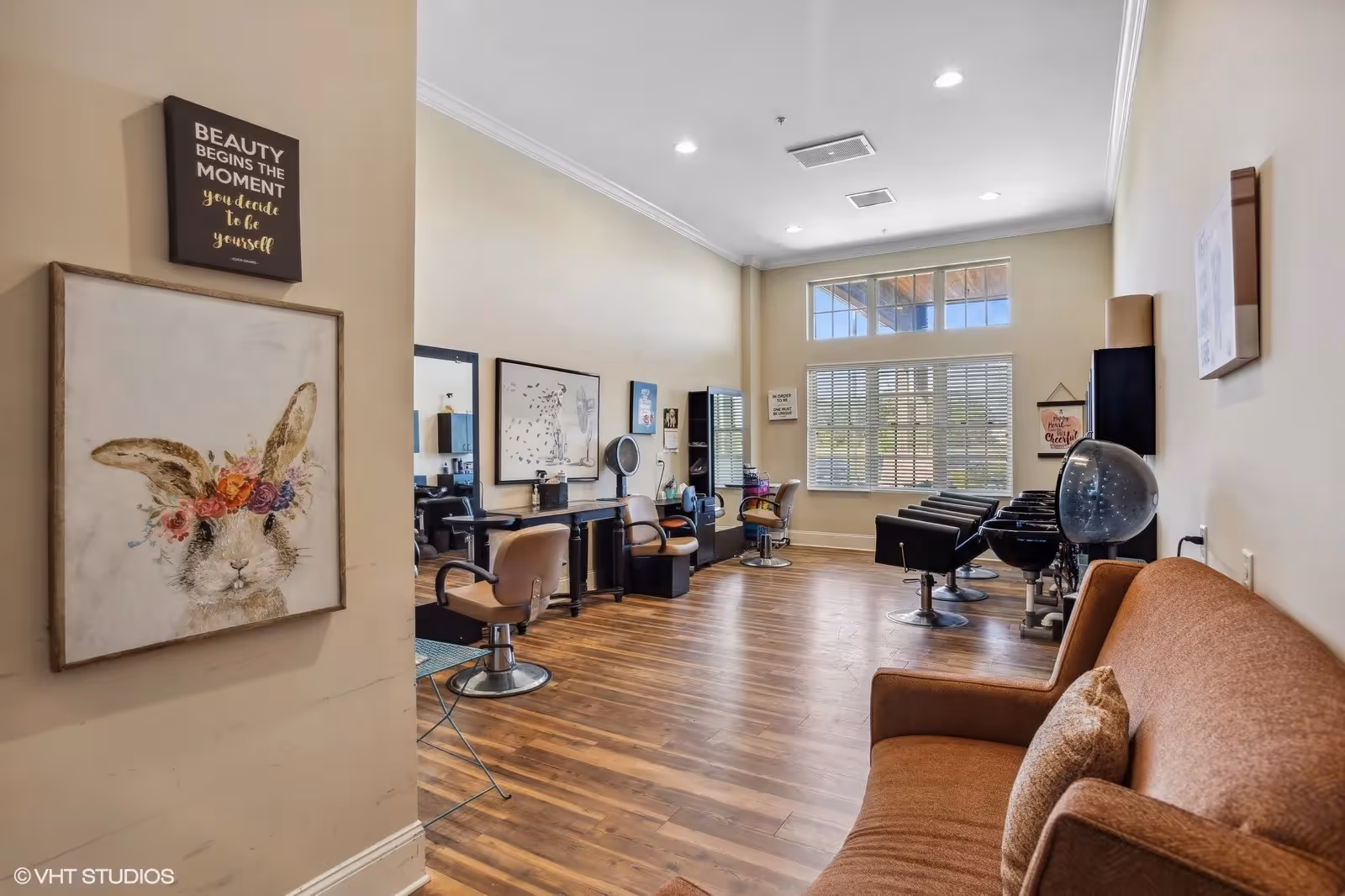 Interior view of a hair salon area with wooden flooring, several salon chairs and hair washing stations along the right wall, a brown couch with a pillow on the right side, and large windows with blinds letting in natural light. Wall decorations include a framed picture of a bunny with a flower crown and a sign that reads 'Beauty begins the moment you decide to be yourself.'