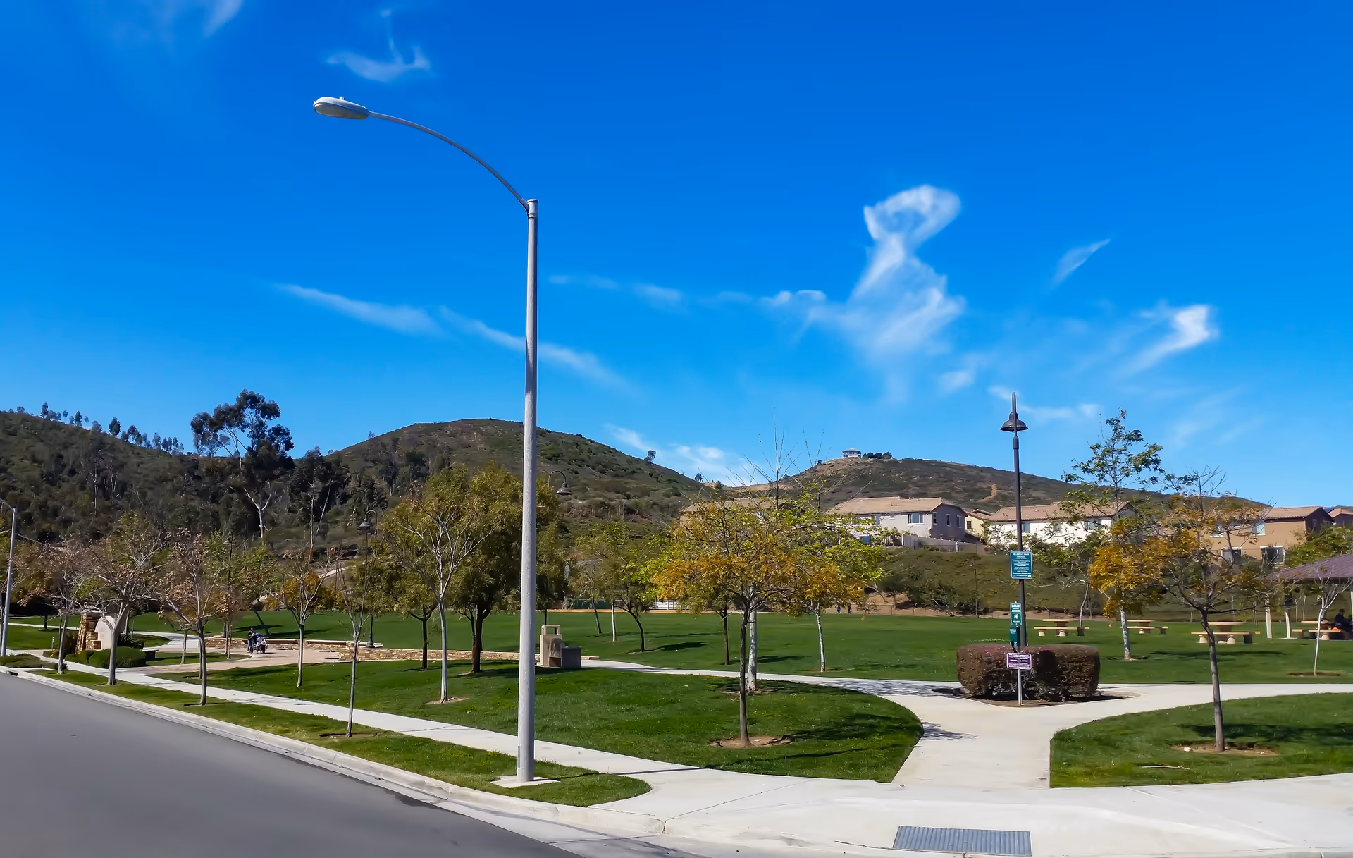A sunny outdoor park area with green grass, young trees, paved walking paths, and a streetlamp. Hills and residential buildings are visible in the background under a clear blue sky with some wispy clouds.