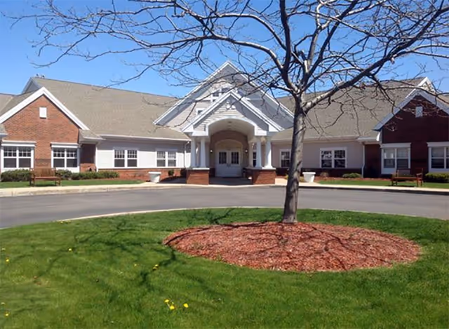 Entrance of a single-story brick-and-siding senior living building with a circular driveway and a tree in the foreground.
