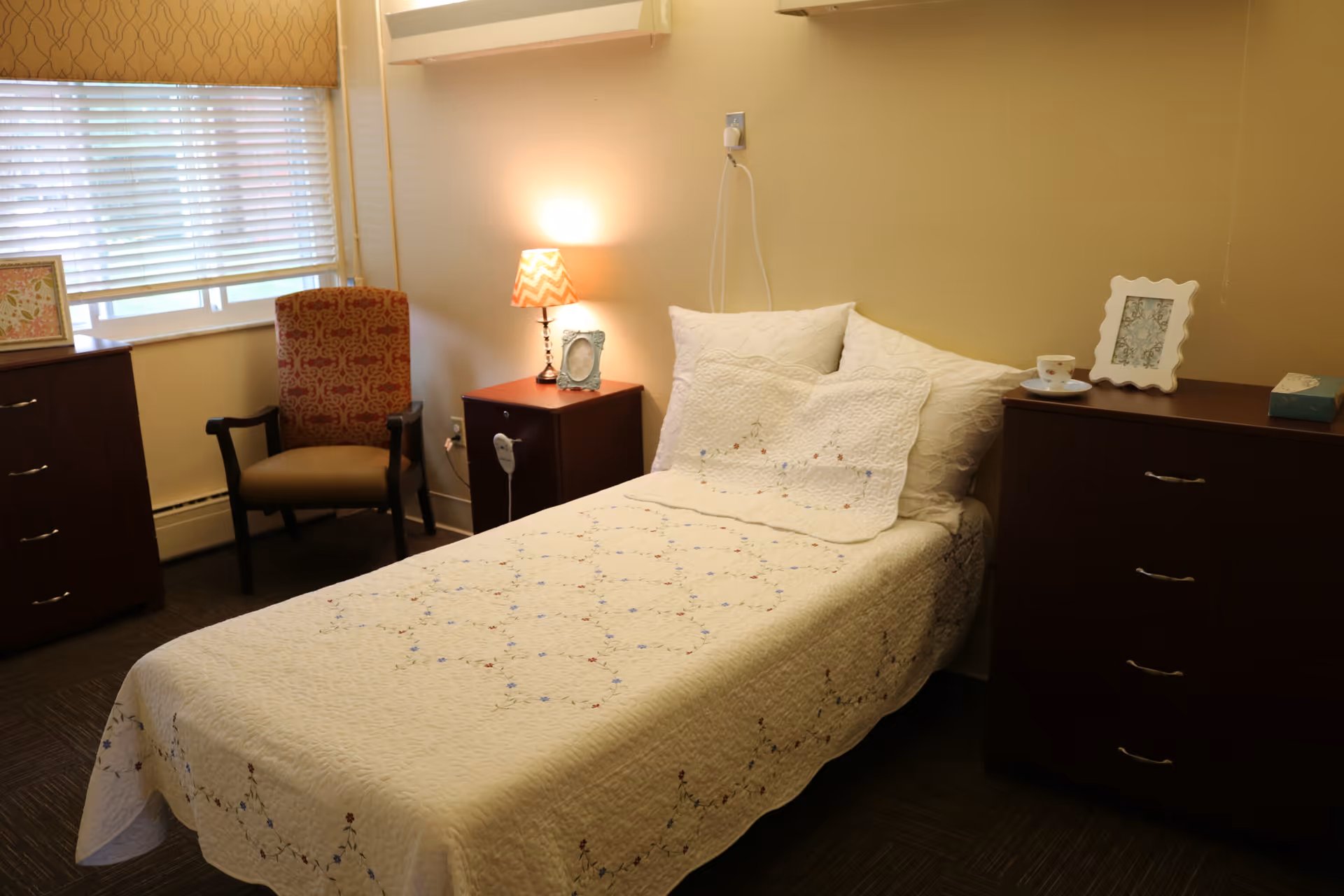 A cozy bedroom with a single bed covered in a white quilt with floral embroidery. There are two wooden dressers on either side of the bed, one with a lamp and a framed photo, and the other with a framed photo, a cup and saucer, and a box of tissues. A patterned armchair is placed near a window with blinds and a valance.
