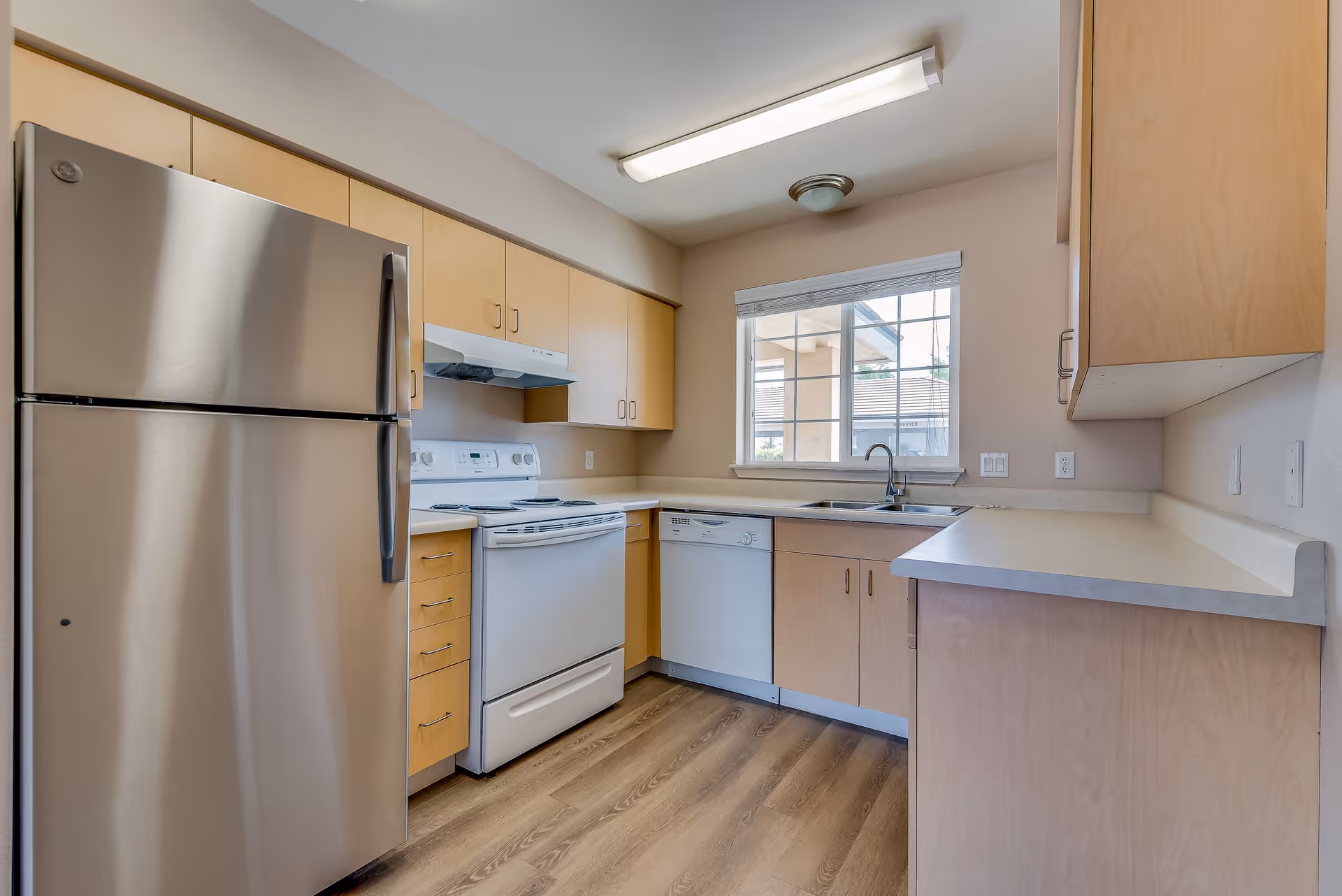 A modern kitchen with light wood cabinets, a stainless steel refrigerator, a white electric stove with an overhead vent, a dishwasher, a sink under a window, and light-colored countertops. The floor is wood-style laminate, and the room is lit by a ceiling light fixture.