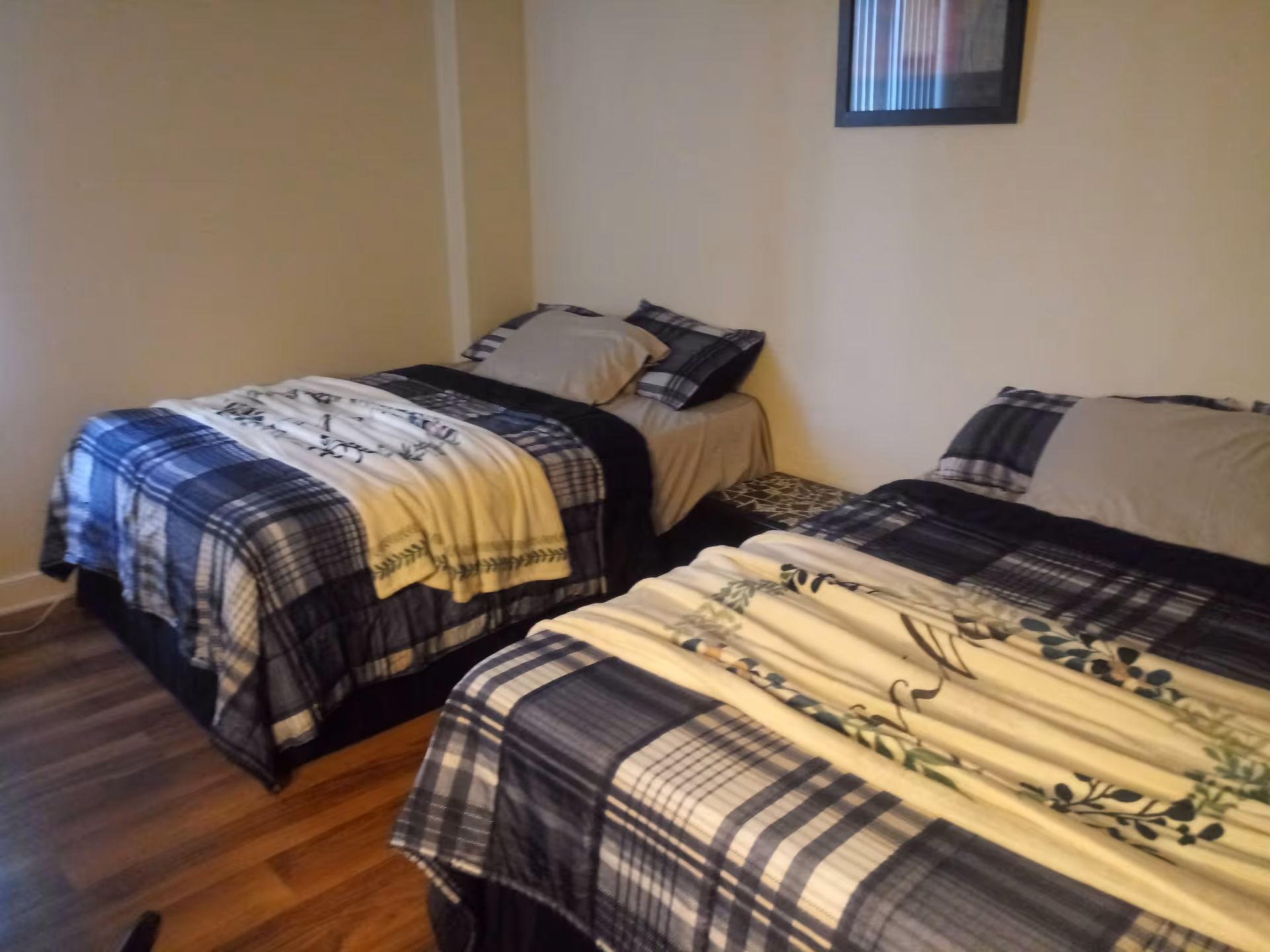 A bedroom with two single beds placed side by side, each with plaid bedding and a light-colored blanket on top. There is a small table between the beds and a framed picture hanging on the wall above.