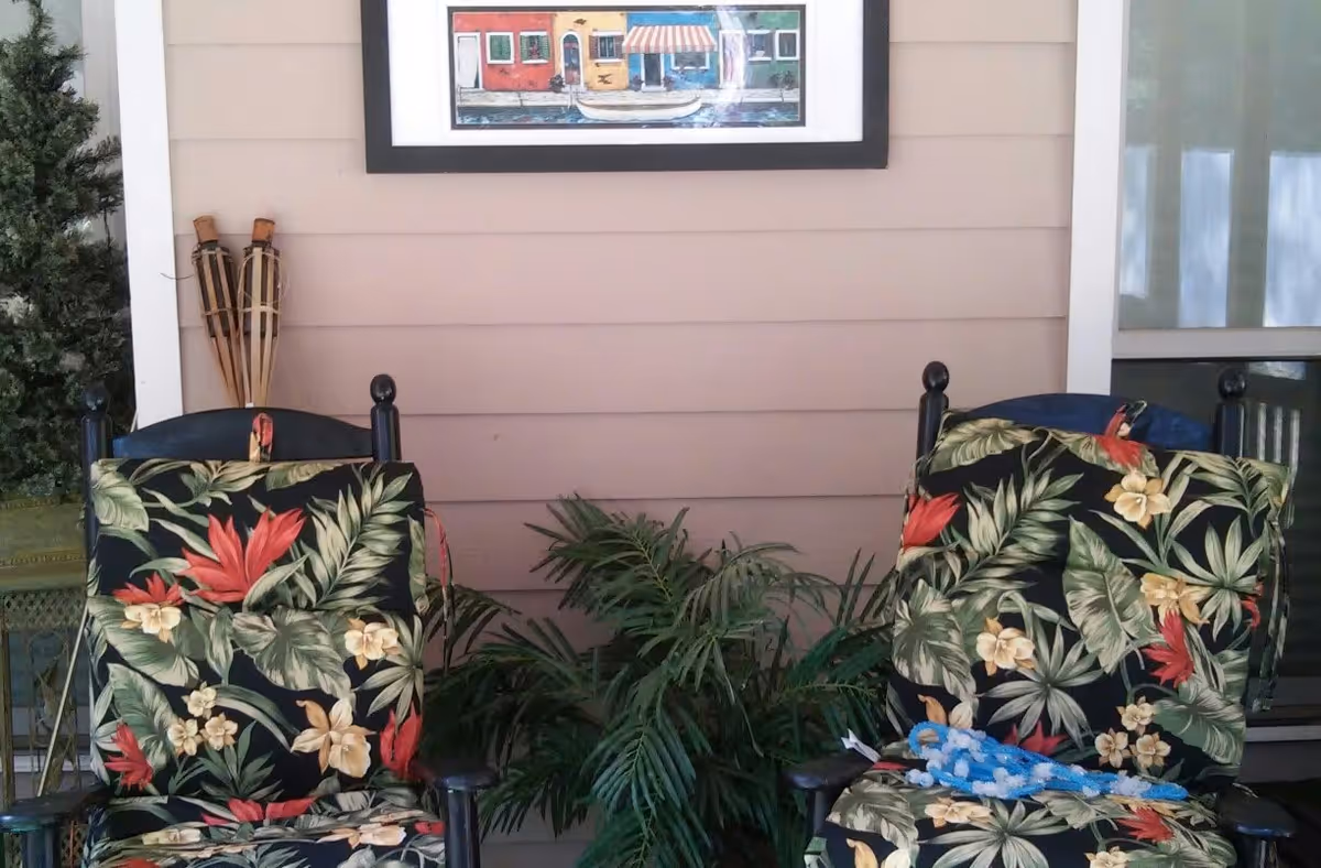 Two black wooden chairs with floral patterned cushions placed on a porch with beige siding. Between the chairs is a green leafy plant. On the wall above the chairs is a framed painting depicting colorful buildings and a boat on water. There are tiki torches leaning against the wall on the left side and a window with white curtains on the right side.