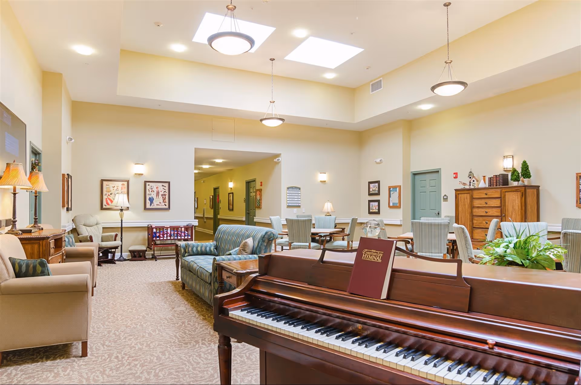 A bright and spacious common area in an assisted living facility featuring a wooden piano with a Christian hymnal on the music stand, several upholstered chairs and sofas, multiple tables with chairs, decorative lamps, framed artwork on the walls, and a skylight ceiling providing natural light.