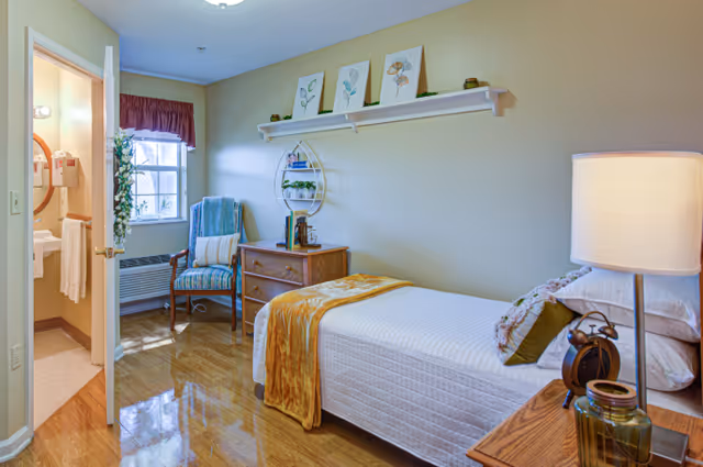 A cozy bedroom with a single bed covered in a white quilt and a mustard yellow throw blanket. There is a wooden nightstand with a lamp, an alarm clock, and decorative items. A wooden dresser with books and plants sits against the wall, which is decorated with three framed botanical prints and a small shelf holding plants. A cushioned chair with a pillow is placed near a window with a maroon valance. The door to a bathroom is open, showing a sink and mirror inside.