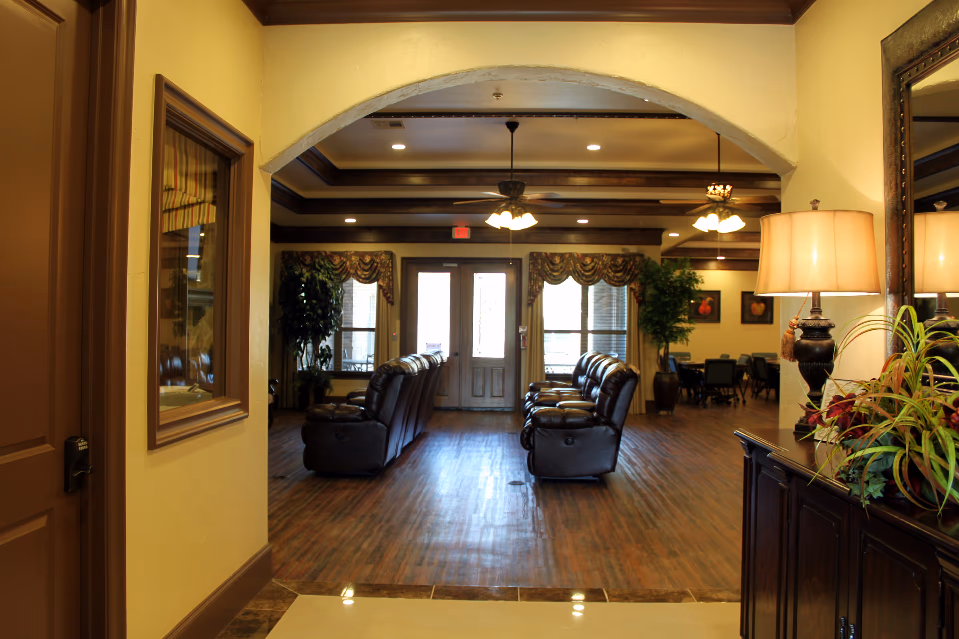 View through an archway into a spacious living room area with two rows of dark leather recliners facing each other. The room has wooden floors, ceiling fans with lights, large windows with decorative curtains, and potted plants. A wooden cabinet with a lamp and plants is visible on the right side.