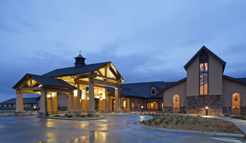 Exterior view of a large senior living facility building at dusk with warm lights illuminating the entrance area and a covered drop-off area supported by columns. The sky is cloudy and the parking lot is wet, reflecting the lights.