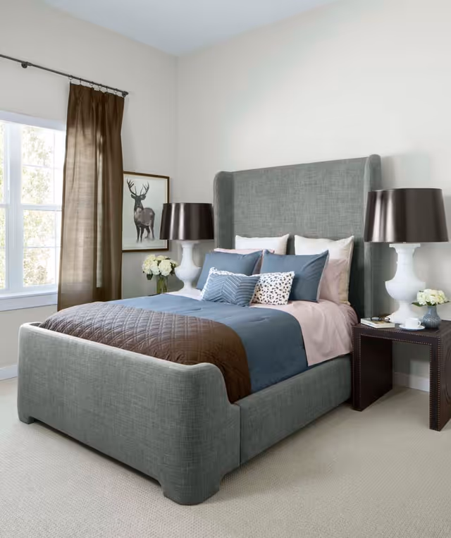 A neatly made bed with a gray upholstered headboard and footboard in a bedroom. The bed has blue and brown bedding with multiple pillows. On either side of the bed are white lamps with dark lampshades on dark bedside tables. A window with brown curtains is on the left wall, and a framed picture of a deer hangs next to the window. There are small vases with white flowers on each bedside table.