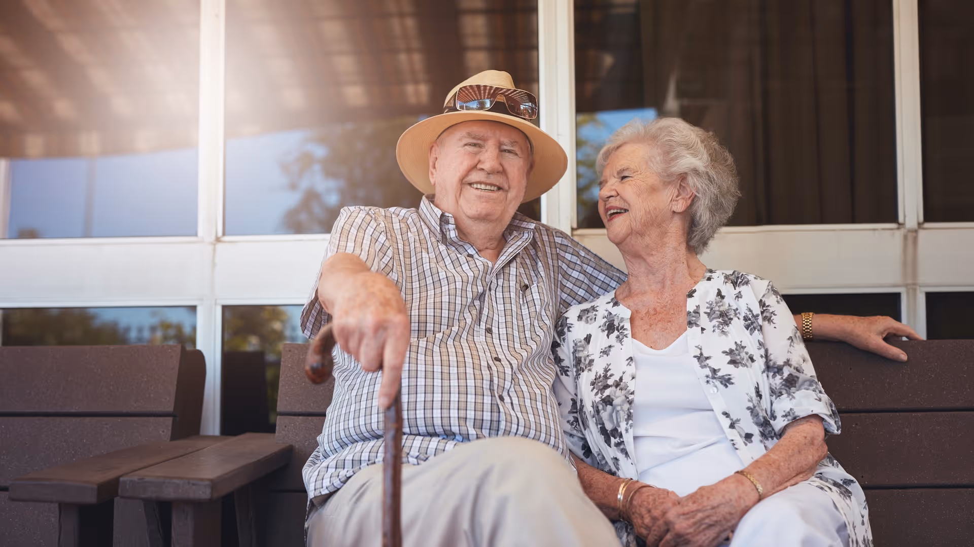 An elderly man wearing a hat and sunglasses on his head sits on a bench next to an elderly woman in a floral cardigan. They are smiling and appear to be enjoying each other's company outside a building with large windows.