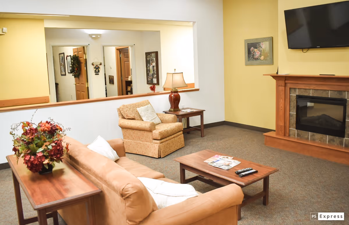 A cozy senior living facility common area with a beige sofa and armchair, a wooden coffee table with magazines and a remote control, a side table with a lamp, a fireplace with a mounted TV above it, and a floral painting on the wall. There is a large mirror reflecting doorways and decorations in the background.