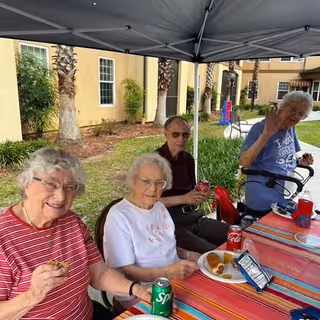 Four elderly individuals sitting at a table outdoors under a canopy, enjoying snacks and drinks including soda cans and chips. They are smiling and appear to be having a pleasant time. The background shows a building with windows and palm trees.