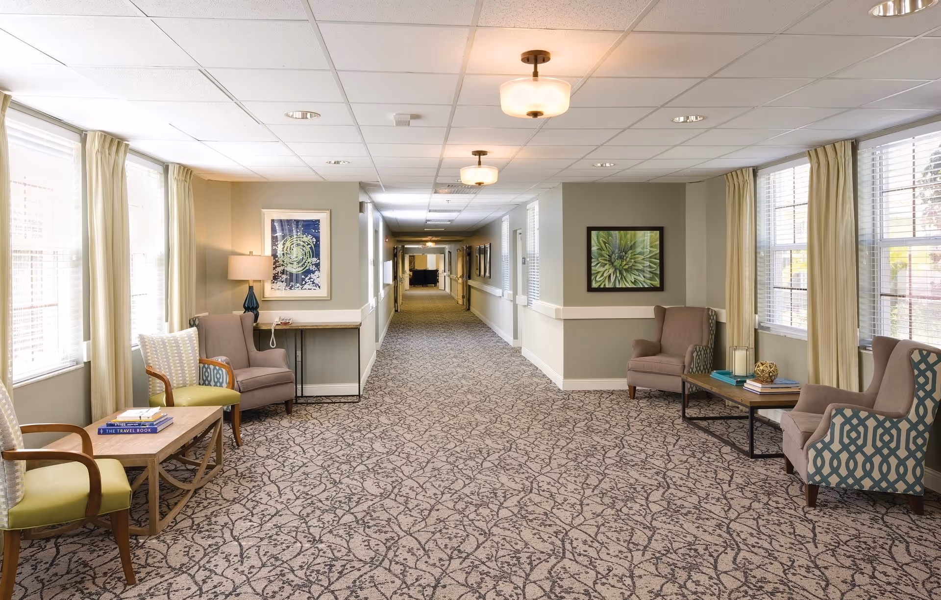 A well-lit hallway in a senior living facility with patterned carpet and beige walls. The hallway is furnished with several armchairs and small tables, some holding books and decorative items. Large windows with cream-colored curtains allow natural light to fill the space. Artwork is hung on the walls, and ceiling lights provide additional illumination.