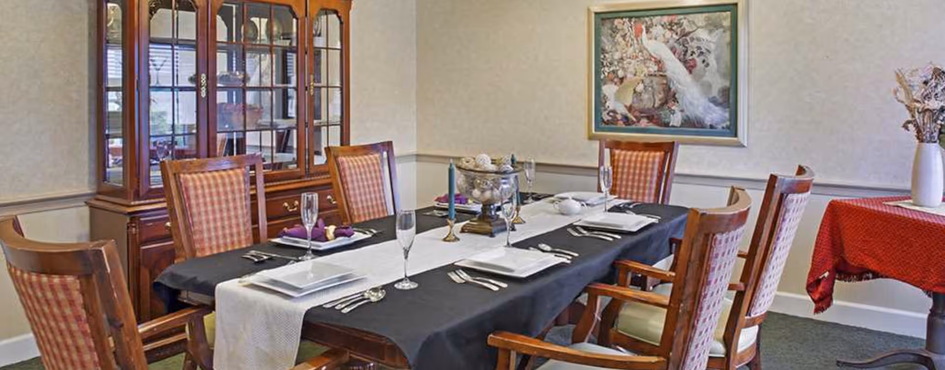 A formal dining room with a long wooden table covered by a black tablecloth and a white runner. The table is set with plates, silverware, glasses, and napkins. Six wooden chairs with cushioned seats surround the table. In the background, there is a wooden china cabinet with glass doors displaying dishes. A framed painting of white peacocks hangs on the wall, and a small table with a red tablecloth and a vase with dried flowers is visible on the right side.