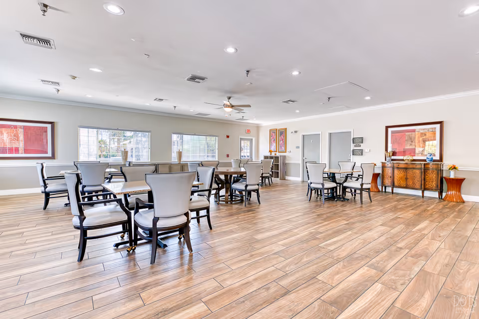 Bright senior living dining area with multiple tables and chairs, wood-look flooring, and framed artwork on the walls.