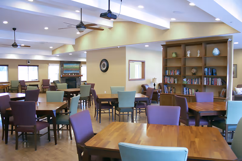 A spacious, well-lit common area with multiple wooden tables and chairs in shades of purple and green. The room features a bookshelf filled with books and decorative items, a wall clock, ceiling fans, and a fireplace in the background. Large windows allow natural light to fill the space.