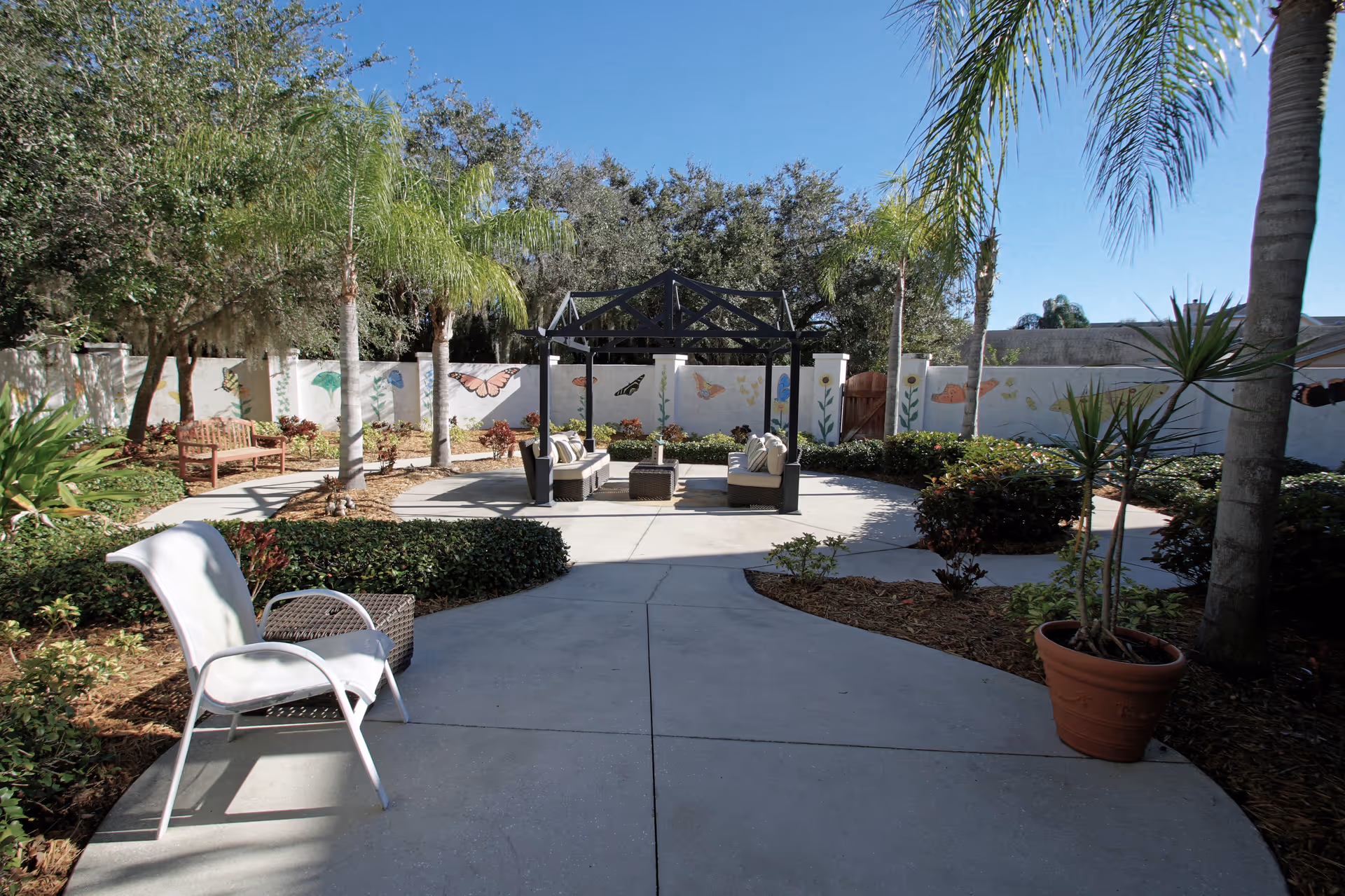 Outdoor courtyard area with a concrete pathway leading to a seating area under a black pergola. The seating area includes cushioned chairs and a small table. Surrounding the pathway and seating area are various plants, palm trees, and bushes. A white wall in the background is decorated with colorful butterfly and flower murals. The sky is clear and blue.