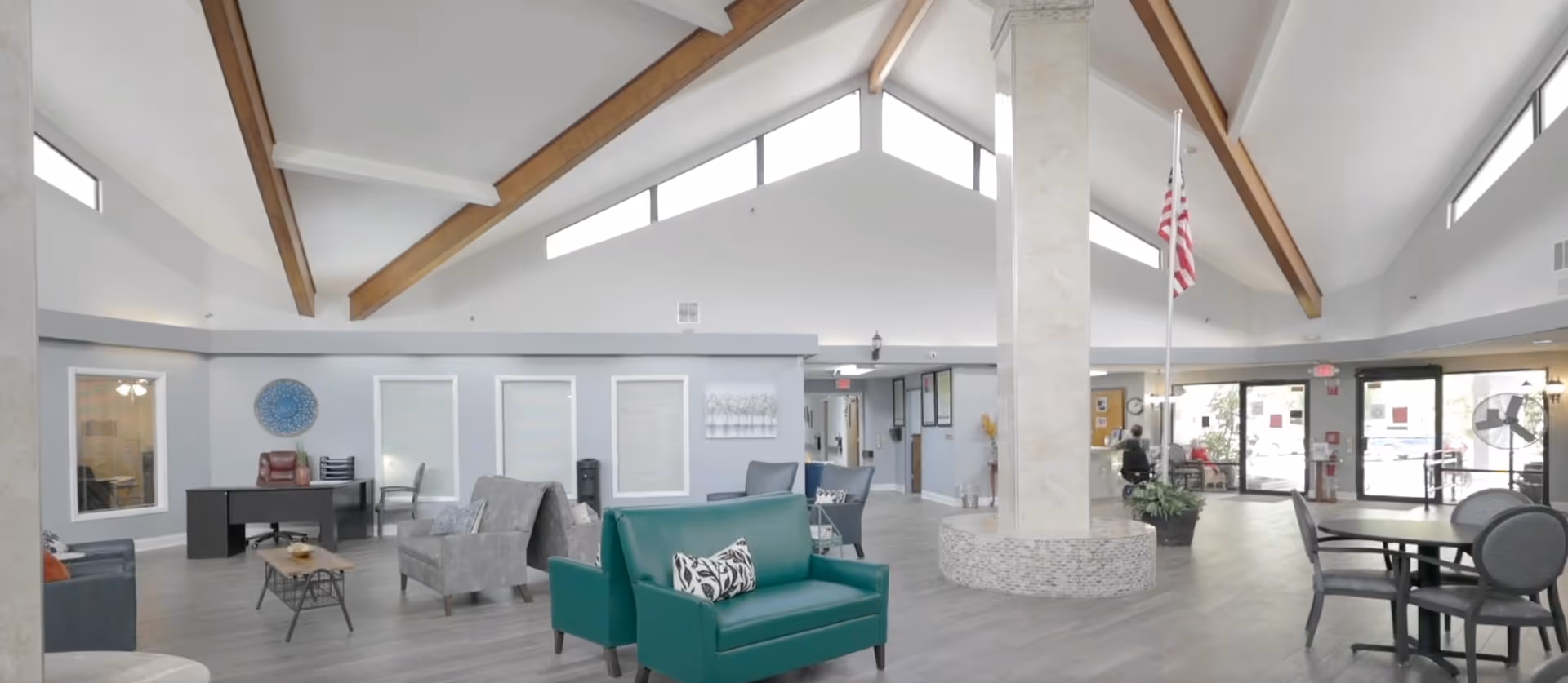 Spacious senior living facility common lounge with vaulted beamed ceiling, seating areas, a central column with an American flag, and glass entry doors.