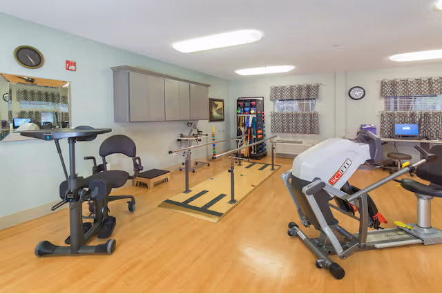 A rehabilitation room with exercise equipment including stationary bikes, parallel bars for walking practice, and a rack with various exercise balls and weights. The room has wooden flooring, light-colored walls, two windows with patterned curtains, a clock on the wall, and a computer workstation in the corner.