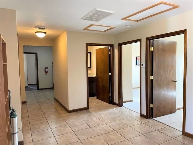 Tiled hallway in an assisted living facility with beige walls and several open wooden doors leading to rooms.