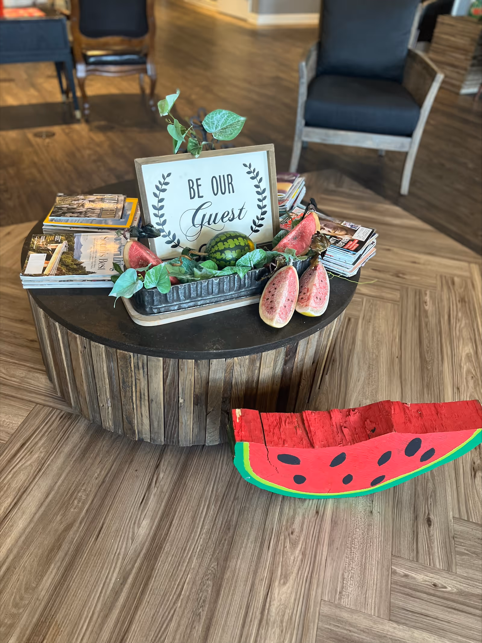 A round wooden coffee table with a decorative tray containing artificial watermelon slices, green leaves, and a small sign that reads 'BE OUR Guest'. The table also holds several stacks of magazines. In the background, there are two wooden chairs with dark cushions on a wood-patterned floor.